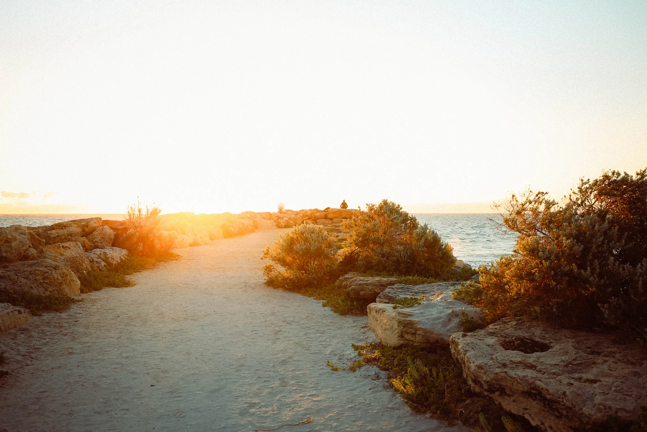 Chemin de sable bordé de rochers et de végétation, menant vers la mer, baigné par la lumière douce d’un lever de soleil.